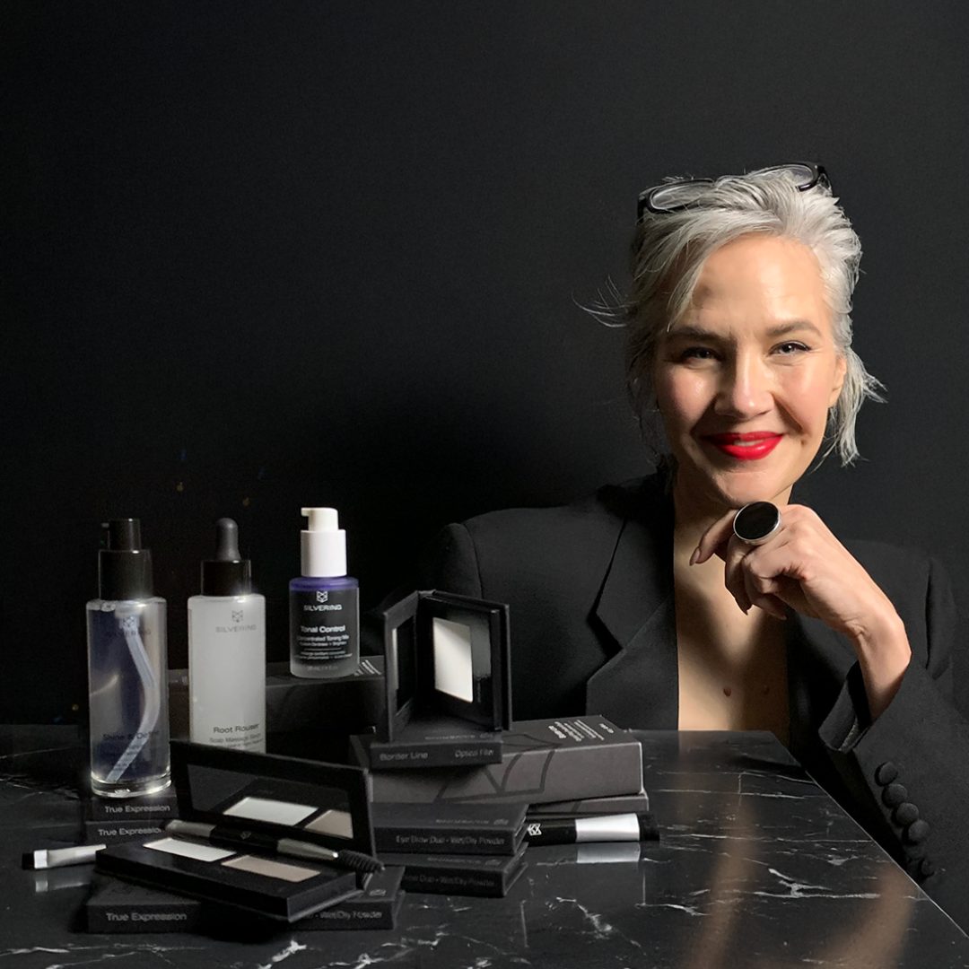 Alexis Eizinas, silver-haired and wearing red lipstick, seated beside Silvering beauty products on a dark marble counter